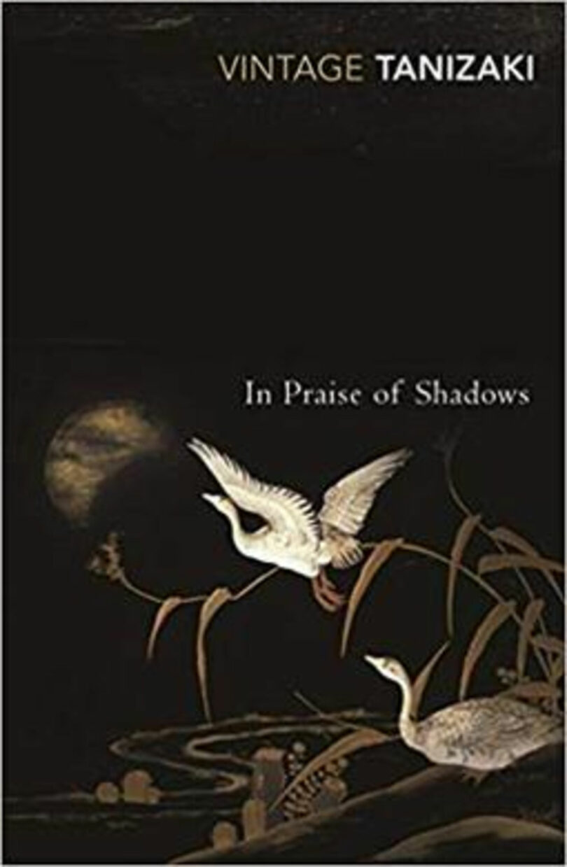 Cover of "In Praise of Shadows" by Tanizaki, featuring two white birds, reeds, a dark background, and a moon or sun partially obscured by clouds with an amber pan of light cast across the scene.
