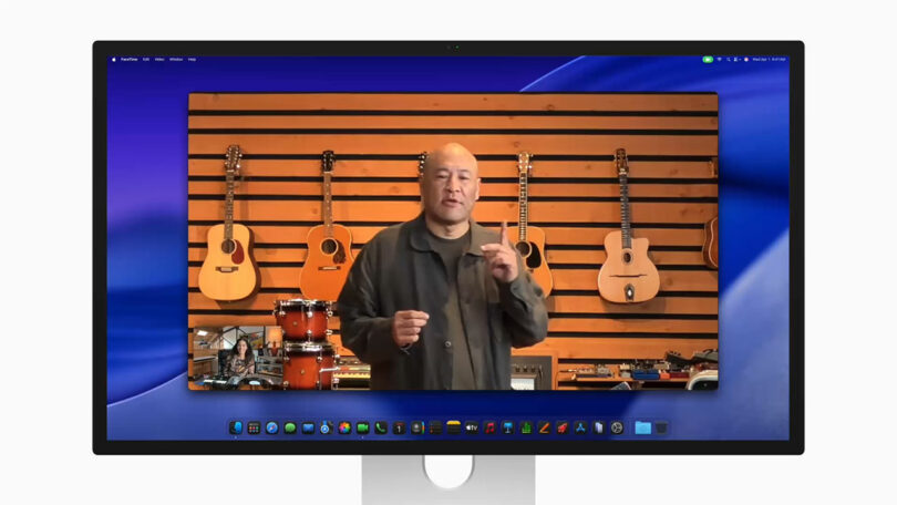 A person stands in front of a wooden slat wall with guitars and percussion instruments, speaking on a video call displayed on a computer monitor.