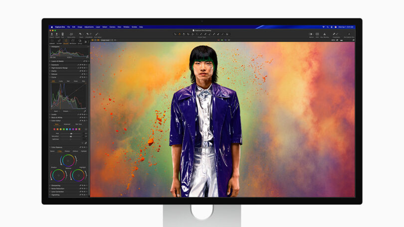 A computer monitor displays a photo editing software with an image of a person in a shiny jacket standing in front of a colorful, powdery background.