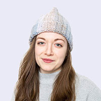 A person with long brown hair is wearing a gray knit hat and a light gray sweater, standing against a plain light purple background.