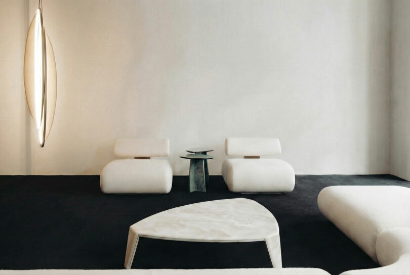 Minimalist living room with two modern white ATRA chairs, a small sculptural side table, a triangular coffee table, and a unique vertical light fixture against a plain white wall and dark floor.