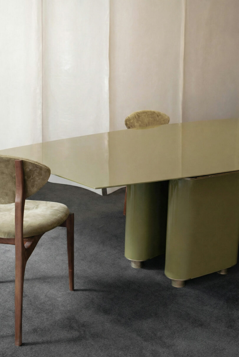 An ATRA modern table with a glossy beige surface and green base stands on a gray carpet, accompanied by two wooden chairs with light upholstered seats and backs.