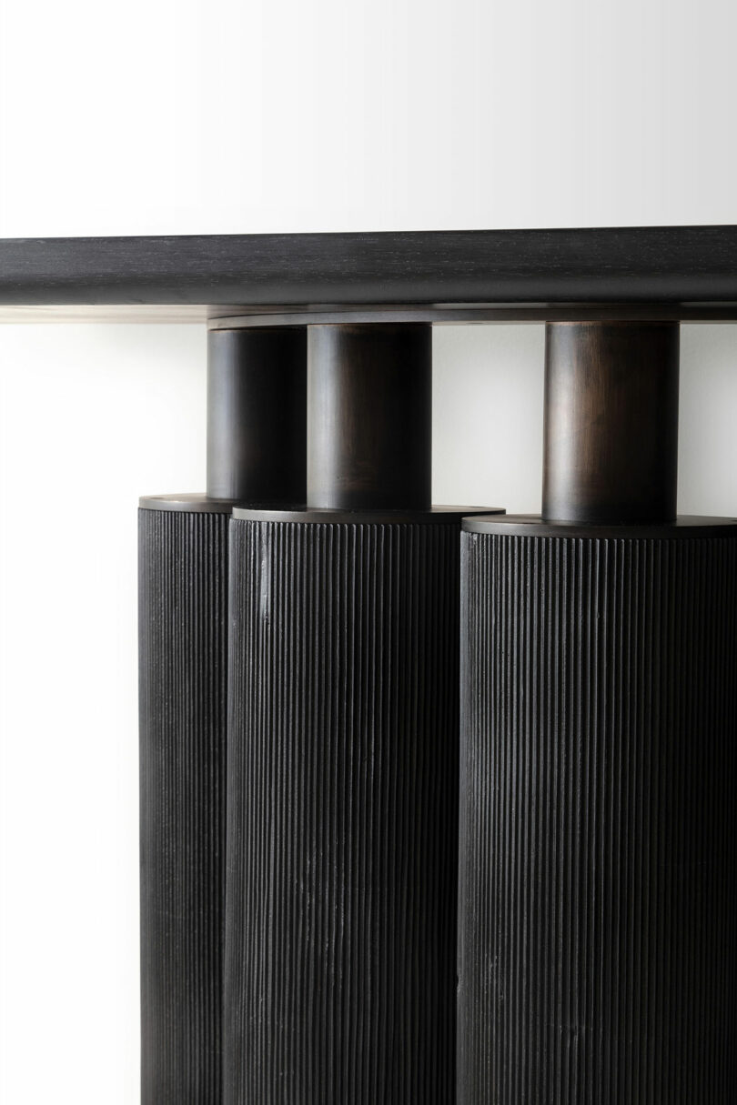 Close-up of a modern black console table by ATRA with three vertically ridged cylindrical legs beneath a smooth, flat tabletop against a white background.