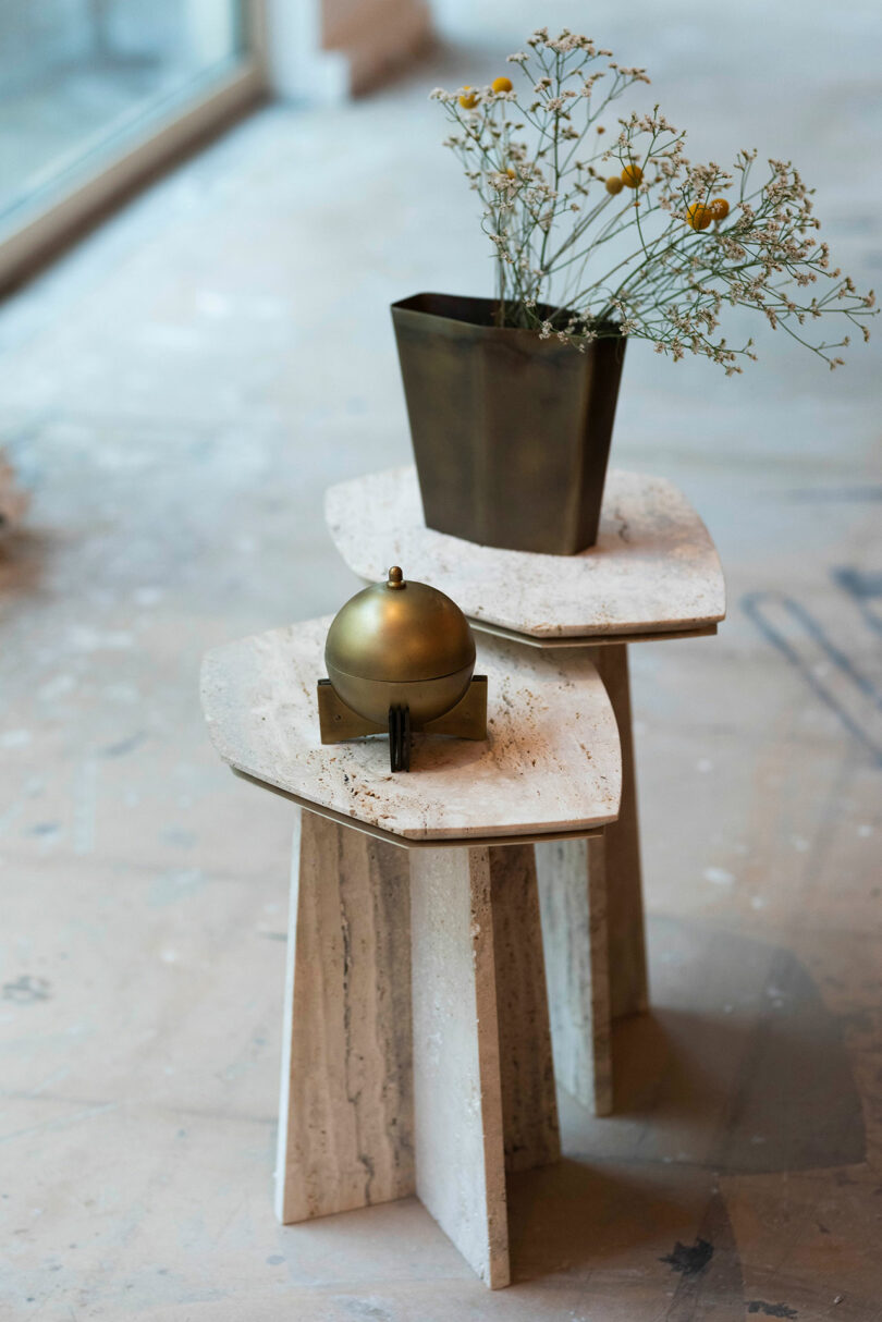Two stacked stone ATRA side tables with a brass spherical object and a brass vase holding dried flowers, placed on a concrete floor near a window.