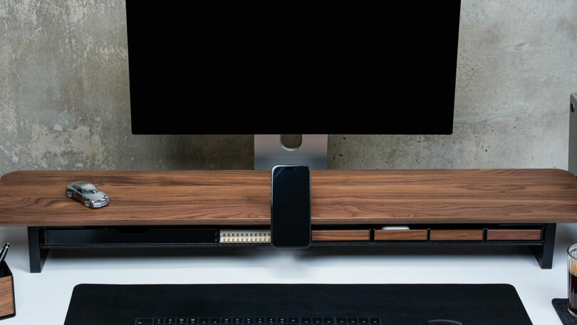 A minimalist desk setup with a Balolo wooden monitor stand, a desktop monitor, a toy car, a smartphone, and neatly organized accessories against a concrete wall.