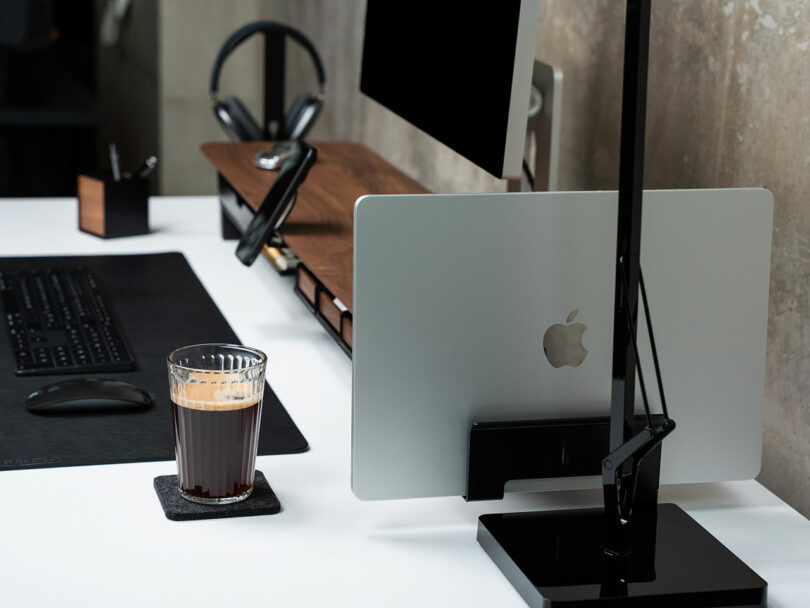 Minimalist desktop setup featuring a monitor, Balolo vertical laptop stand holding a closed MacBook, keyboard, mouse, headphones, and a glass of coffee on a coaster.