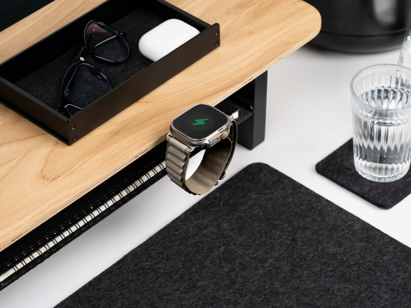 A smartwatch is charging on the edge of a wooden Balolo desk next to a drawer containing sunglasses and earbuds, with a glass of water and a felt desk mat nearby.