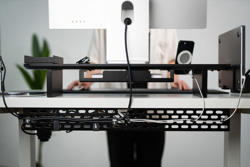 Person standing at a modern desk with a Balolo monitor stand, laptop, and organized cable management tray visible underneath.