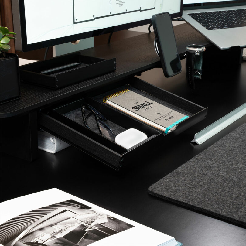 A close-up of a modern desk setup with a Balolo computer monitor stand, phone holder, laptop, camera, open drawer with notebook and accessories, and an open magazine.
