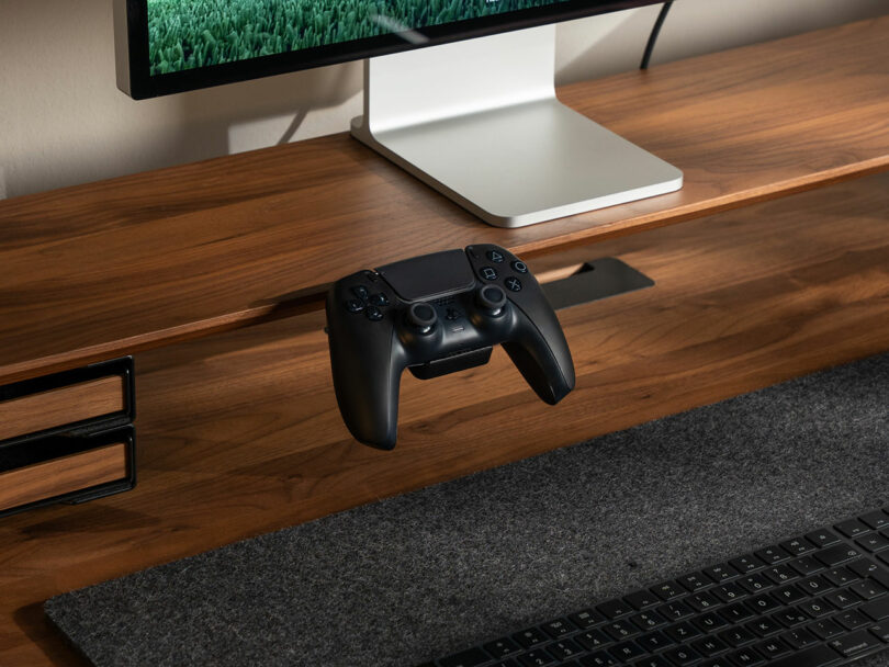 A black game controller is mounted on a Balolo wooden desk under a computer monitor, with a keyboard and a gray desk mat below.