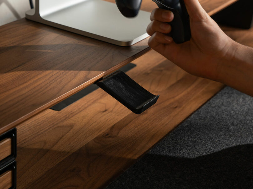 A hand places a black game controller onto a slim, angled Balolo holder attached under a wooden desk.