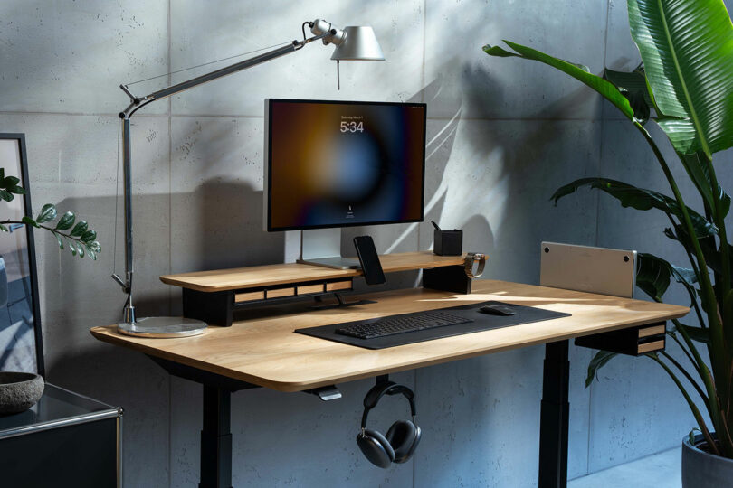 A modern Balolo desk setup featuring a large monitor, keyboard, mouse, lamp, headphones, and potted plants in a well-lit workspace with a concrete wall background.