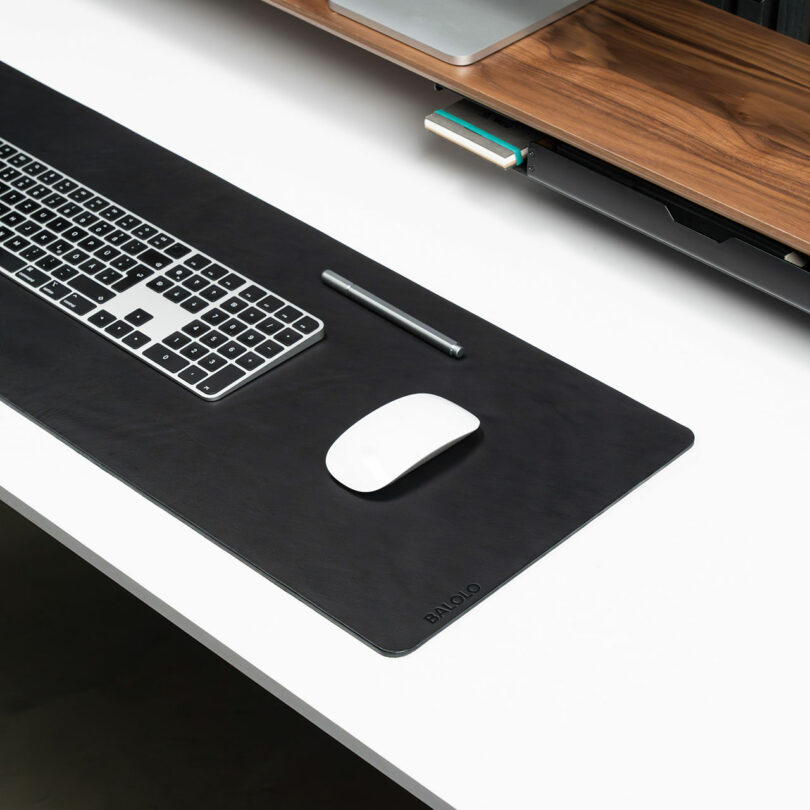 A black desk mat with a keyboard, stylus, and white wireless mouse is arranged neatly on a white desk below a wooden Balolo shelf.