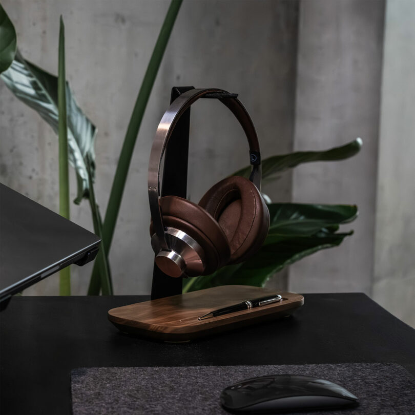 Brown Balolo over-ear headphones rest on a wooden stand next to a pen, with a mouse and laptop partially visible on a black desk. Large green leaves are in the background.