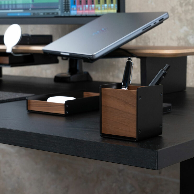A modern desk setup featuring a sleek Balolo dark wooden organizer holding pens, a small tray with essentials, and a laptop on a stand in the background.