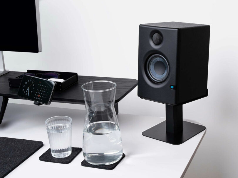 A black Balolo speaker on a stand sits next to a glass and a carafe of water on coasters, with part of a monitor and an organizer visible on a white desk.