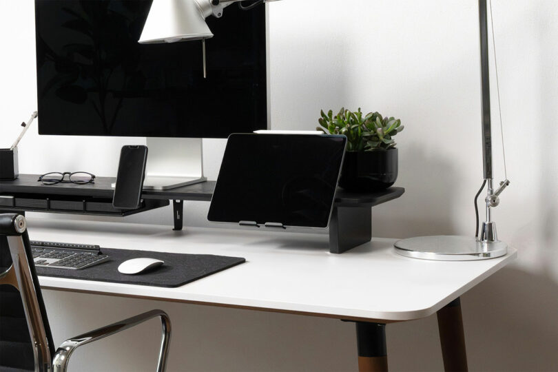 A modern desk setup featuring a Balolo monitor stand, keyboard, mouse, tablet on a stand, smartphone, potted plant, and two desk lamps arranged neatly on a white surface.