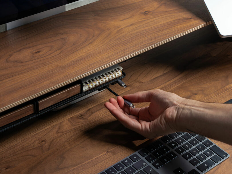 A hand pulls out a Balolo black pen from a built-in compartment beneath a wooden desk shelf, next to a spiral notebook and a keyboard.