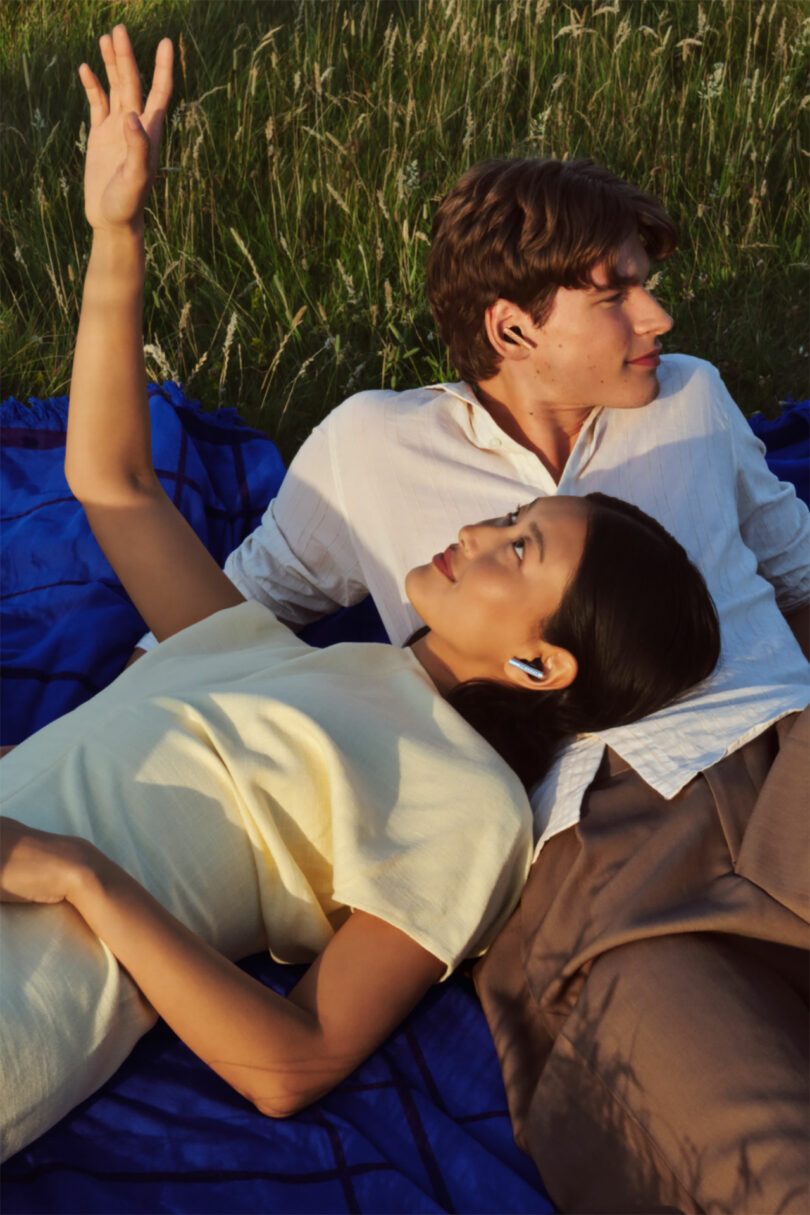 A woman wearing wireless earbuds lies on a blue blanket in the grass with her head on a man's lap, raising her hand while he looks into the distance.