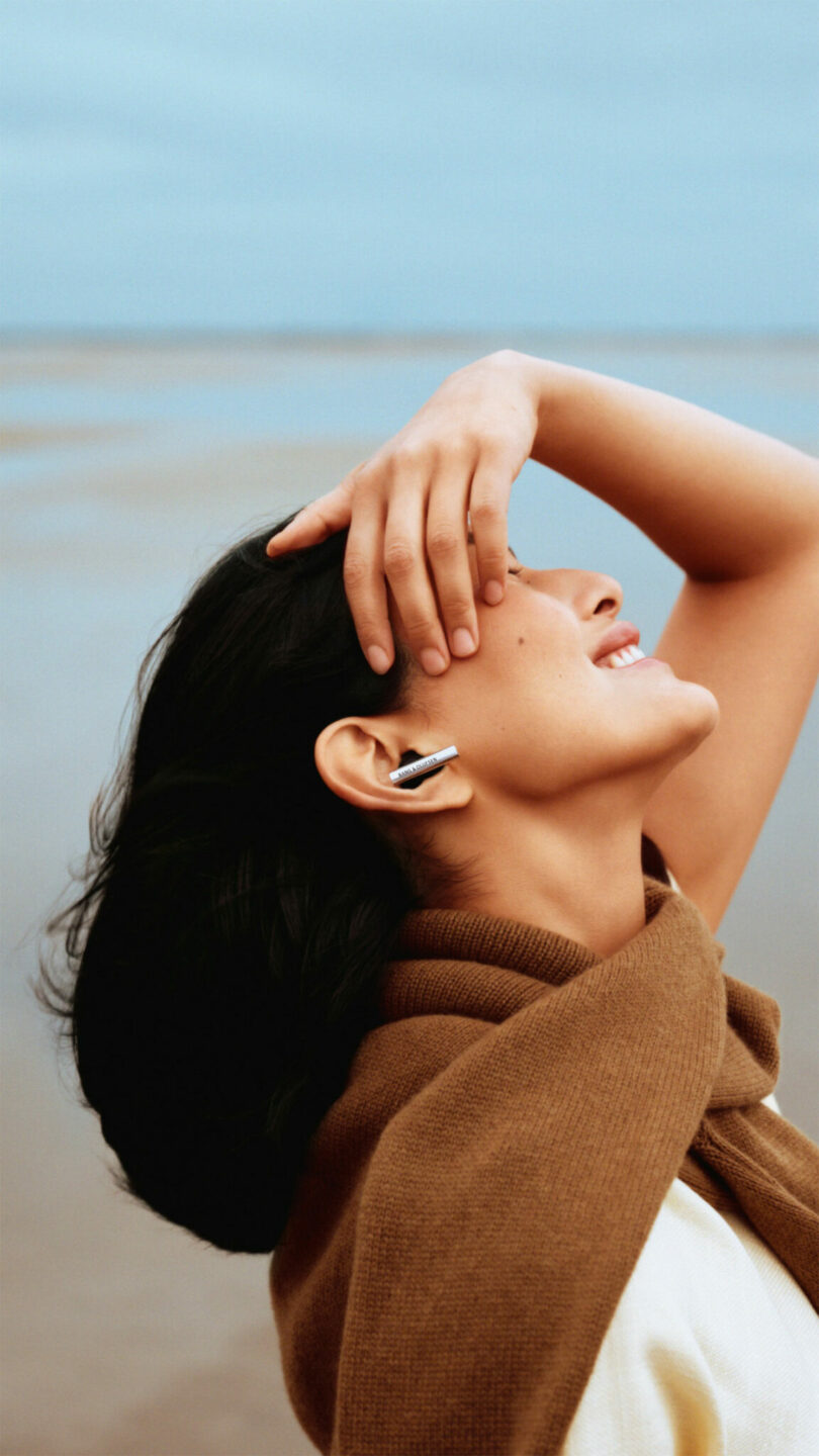 A person wearing a brown scarf stands outdoors, tilting their head back and smiling with one hand resting on their forehead. The background shows a calm, open landscape.