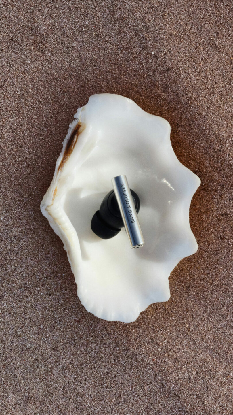 A metallic wireless earbud rests inside a white seashell on brown sand.