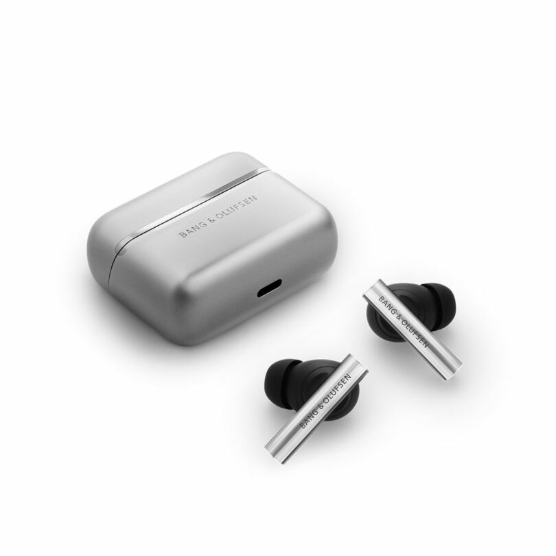 A pair of silver Bang & Olufsen wireless earbuds with black tips are shown next to their matching rectangular charging case.