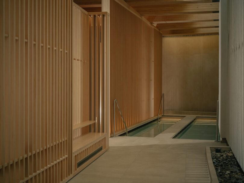 Indoor pool area with wooden slat walls, a narrow rectangular pool, metal handrails, and a stone border along one side. Natural light filters through the ceiling.