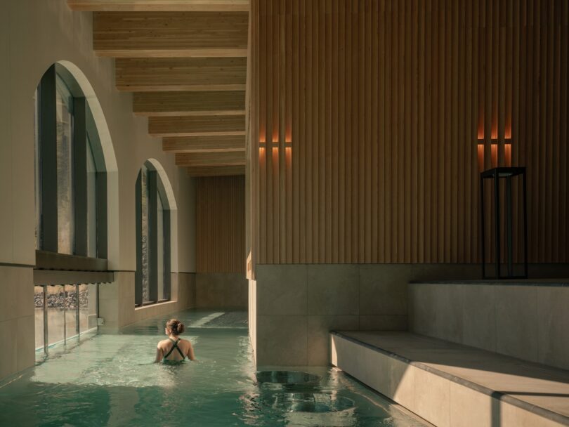 A person swims alone in an indoor pool with large arched windows and wood-paneled walls illuminated by soft wall lights.