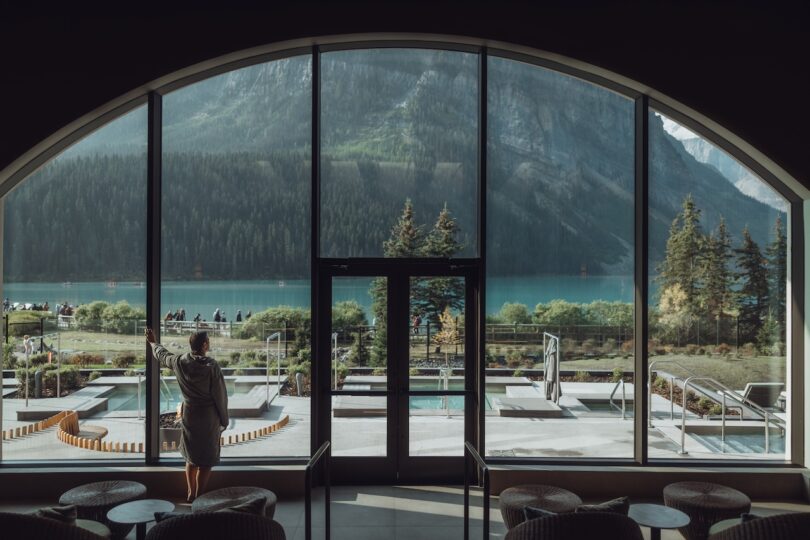 A person in a robe stands indoors by large glass windows, overlooking a patio, a lake, and mountains surrounded by trees.