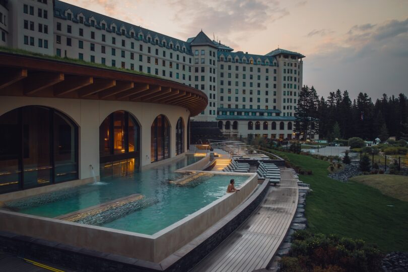 Woman sitting in an outdoor infinity pool at a large hotel resort during sunset, with lounge chairs and landscaped gardens visible.