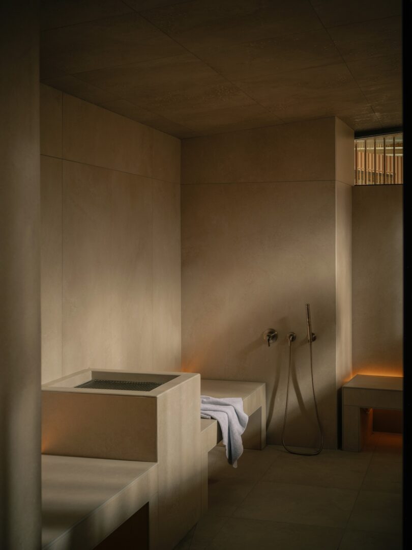 Minimalist bathroom with beige walls, built-in bench, towel, handheld shower, and soft ambient lighting.