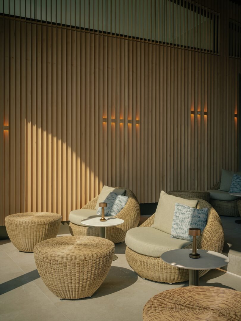 A modern lounge area with wicker chairs and round ottomans, light gray cushions with patterned pillows, and wood slat wall paneling in warm natural light.