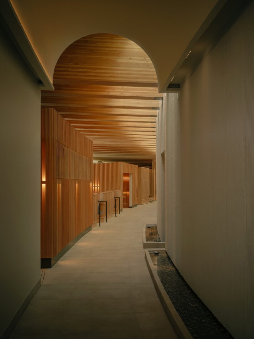 A modern hallway features wood paneling, a curved ceiling, and recessed lighting, with a narrow water feature running alongside the right wall.