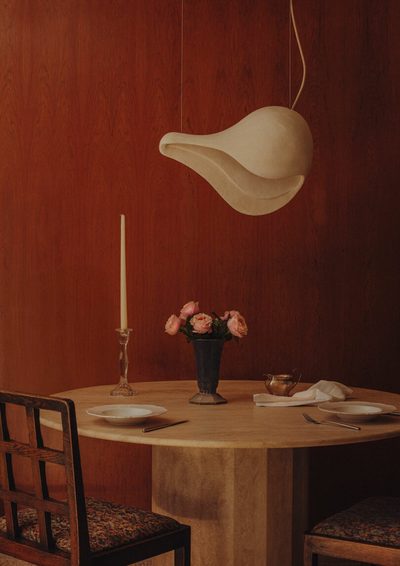 A round table set for two with plates, cutlery, a vase of pink roses, and a single tall candle, under a sculptural white pendant light against a wooden wall.