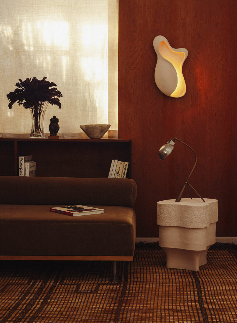 A cozy living room corner with a brown sofa, a sculptural white side table, a metallic desk lamp, and a wavy wall sconce on wood-paneled walls. A shelf holds vases and books.