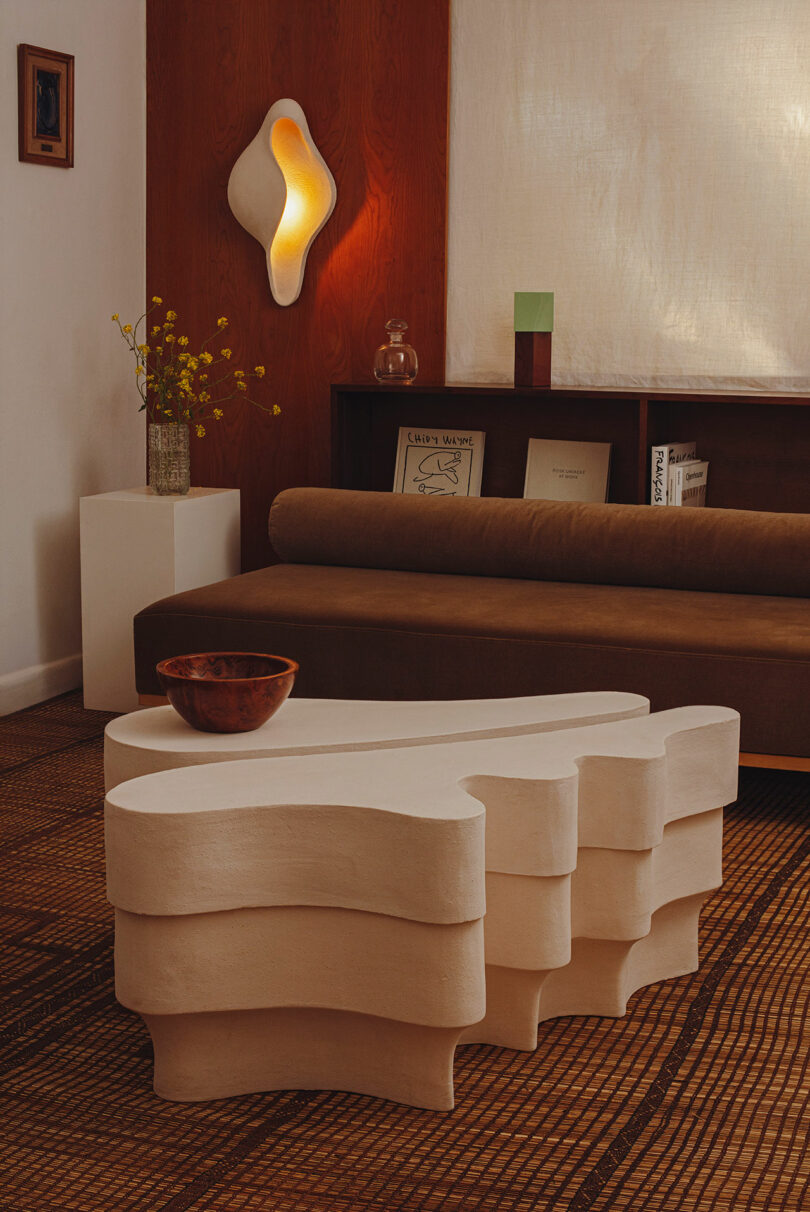 Modern living room with a sculptural, layered white coffee table, a brown sofa, a wooden bowl, wall art, and a glowing organic-shaped wall light. Books and decor are on a shelf behind the sofa.