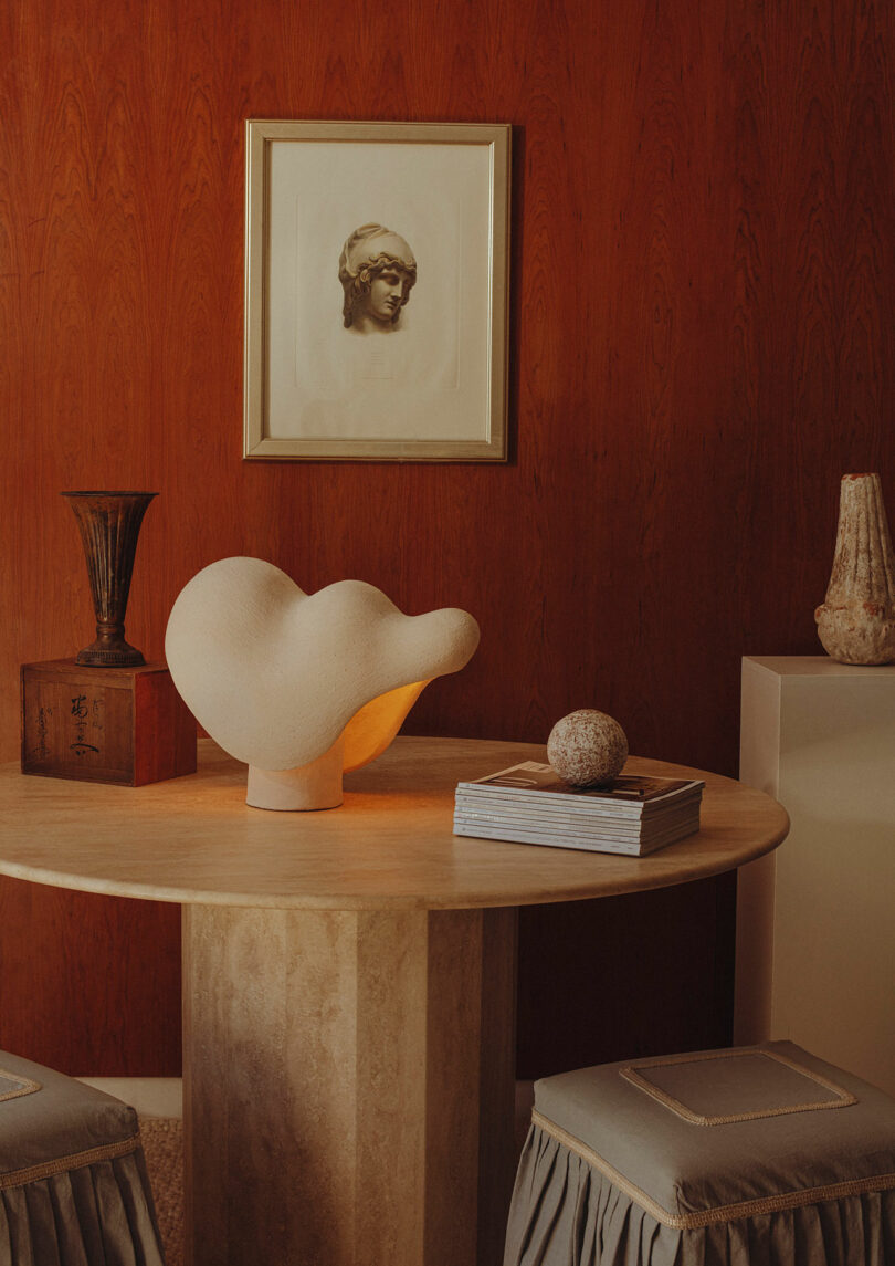 Round beige table with sculptural lamp, books, and decorative objects against a wood-paneled wall with framed art. Two upholstered stools are partially visible.