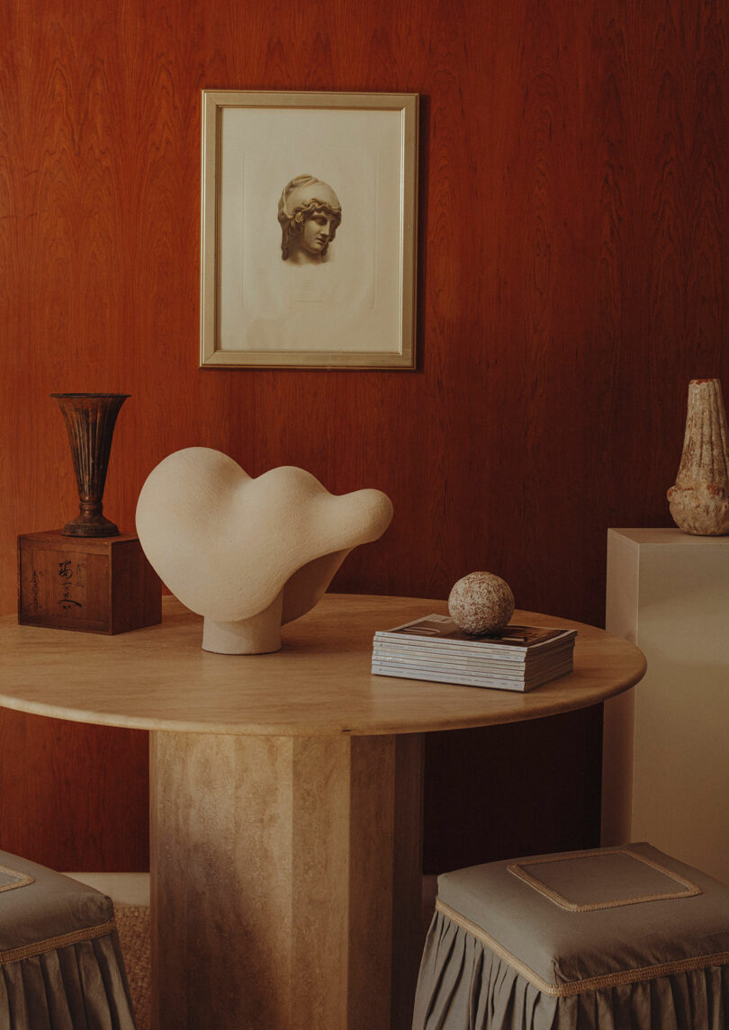Round table with abstract sculpture, stacked books, a sphere, and decorative objects against a wood-paneled wall with framed artwork above. Two stools are placed around the table.