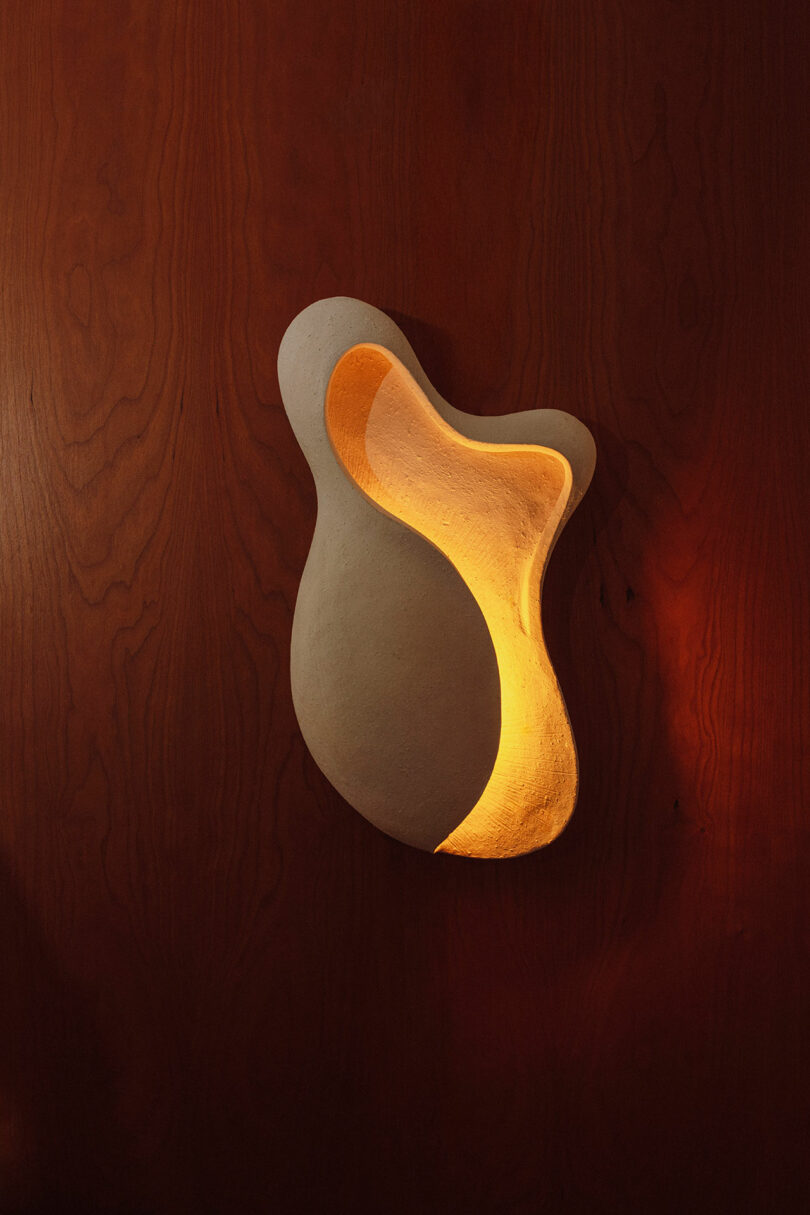 An abstract, white sculptural wall sconce emits warm yellow light against a smooth, reddish-brown wooden wall.