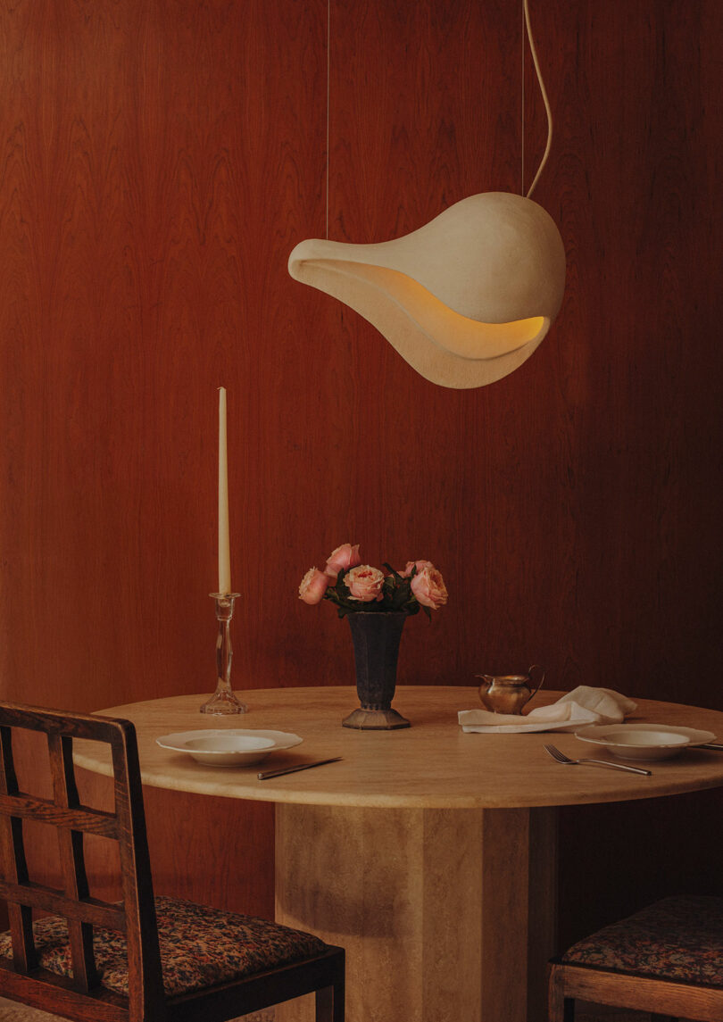 A round table set for two with plates, cutlery, a vase of pink flowers, a candlestick, and a unique white pendant lamp against a wooden wall.