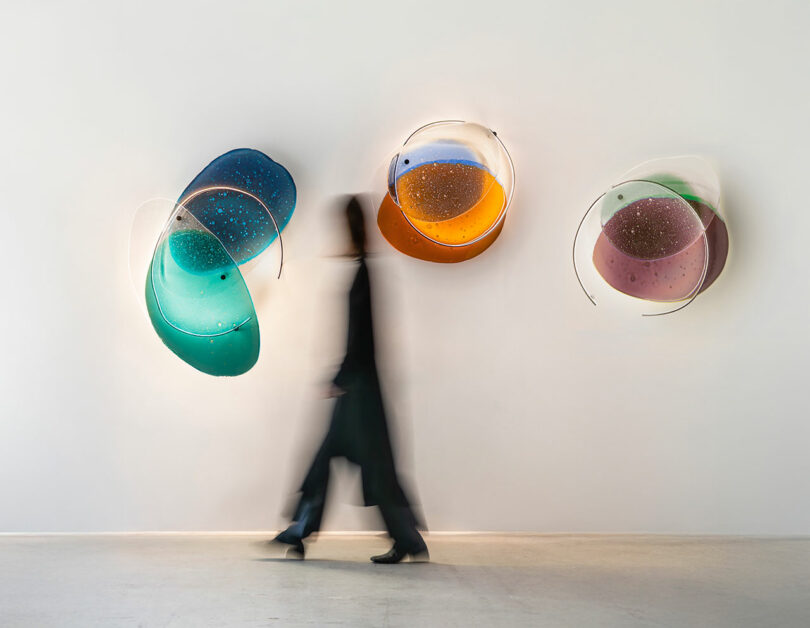 A person in motion walks past three abstract, colorful wall art pieces with overlapping shapes and speckled patterns on a white gallery wall.