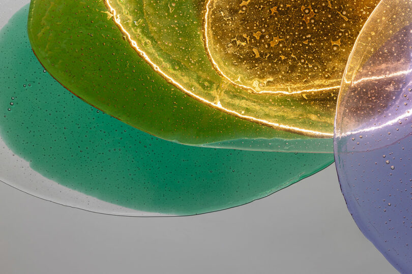 Close-up of overlapping translucent discs in green, yellow, and purple hues, with fine bubbles and textured surfaces, set against a neutral background.