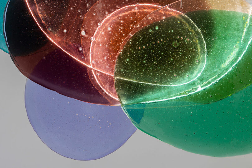 Overlapping translucent colored glass discs in purple, red, orange, and green with visible bubbles, set against a plain background.
