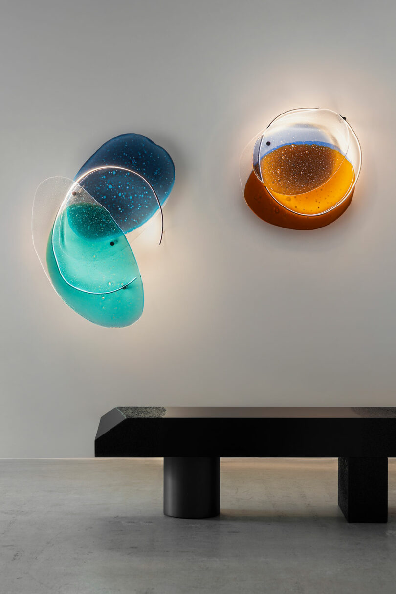 Two abstract, colorful glass wall sculptures—one blue-green and one amber—are mounted on a gray wall above a modern black bench in a minimalist setting.