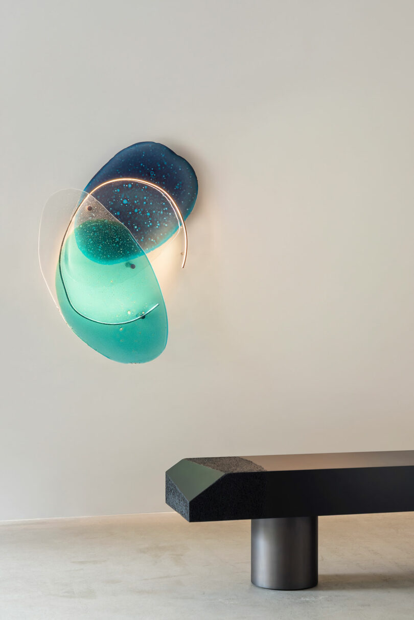 A blue glass sconce hangs on a white wall, and there is a bench in the foreground.