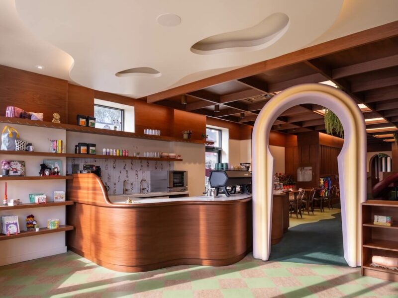 Modern café interior with wooden counters, open shelves displaying items, a coffee machine, and a large decorative archway leading to a seating area with tables and chairs.