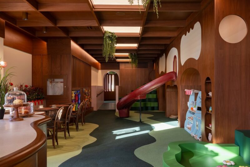 A modern indoor playroom with wooden walls, a red slide, children's toys, seating area, and hanging plants under skylights.