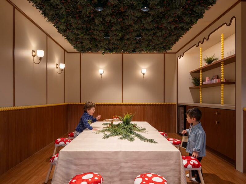 Two young children stand by a table with a beige tablecloth and plants, surrounded by mushroom-shaped stools in a whimsical room with a leafy ceiling.