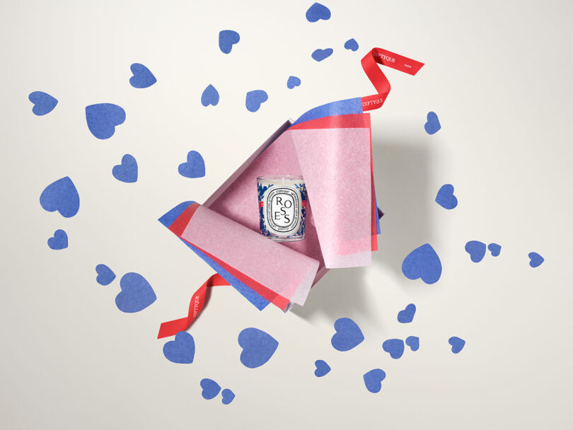 An open pink and red gift box with a Diptyque Roses candle inside, surrounded by scattered blue paper heart cutouts and a red ribbon on a light background, perfect for celebrating Valentine's Day.