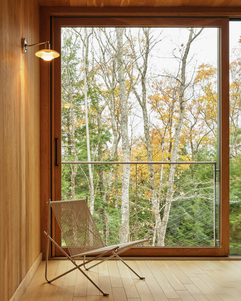 A minimalist room with wooden walls and floor features a Design Within Reach metal chair near a large window overlooking leafless trees and autumn foliage outside.